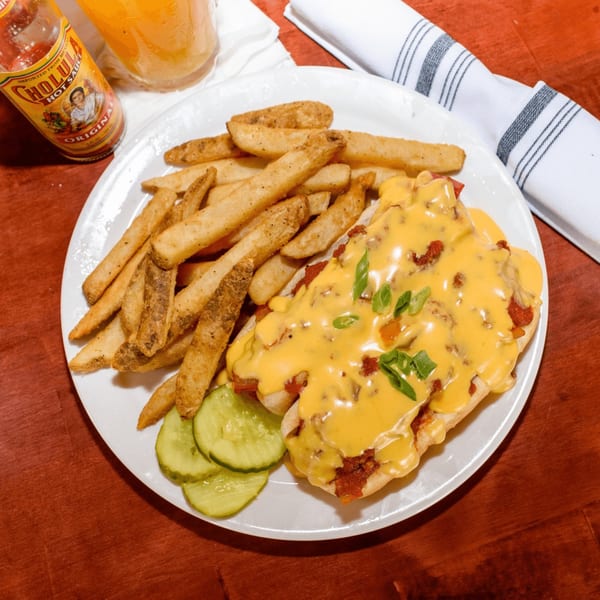 A plate of cheesy chili dogs with fries and pickles, with a bottle of hot sauce.