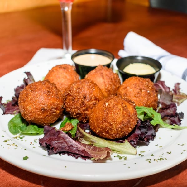 Six golden-brown fried balls, possibly hush puppies, on a bed of mixed greens with two dipping sauces.