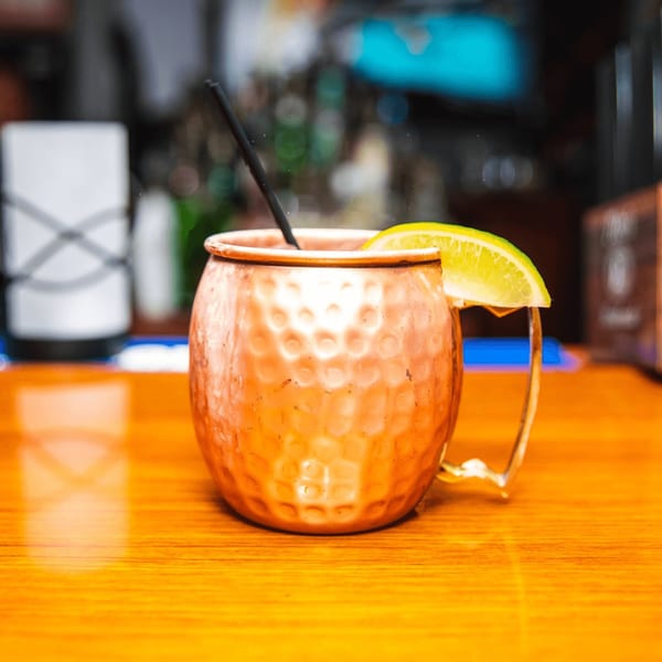 A Moscow Mule in a hammered copper mug with a lime wedge and black straw on a wooden bar.