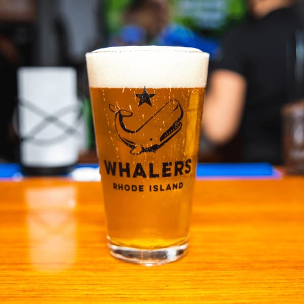 A pint glass of Whalers Rhode Island beer with a foamy head sits on a wooden bar.