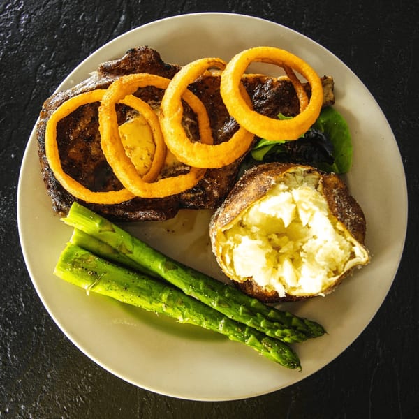 Grilled steak topped with onion rings, served with a baked potato and asparagus on a white plate.