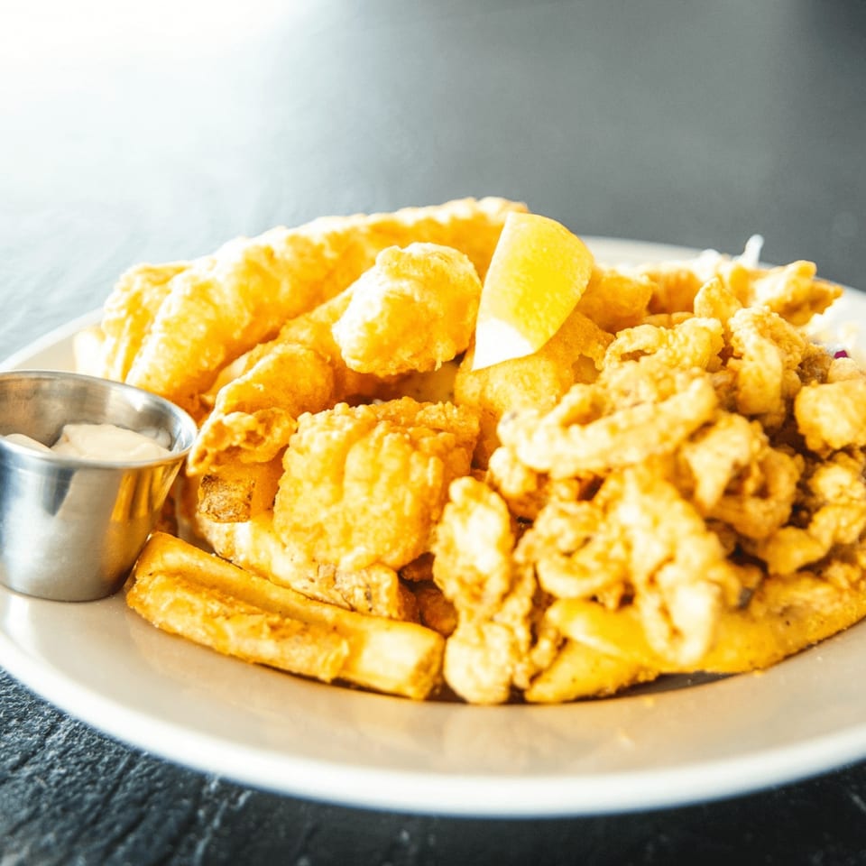 A platter of golden-fried fish, calamari, and fries with a side of tartar sauce and lemon.